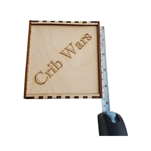 Wooden box with 'Crib Wars' engraving next to a measuring tape on a white background